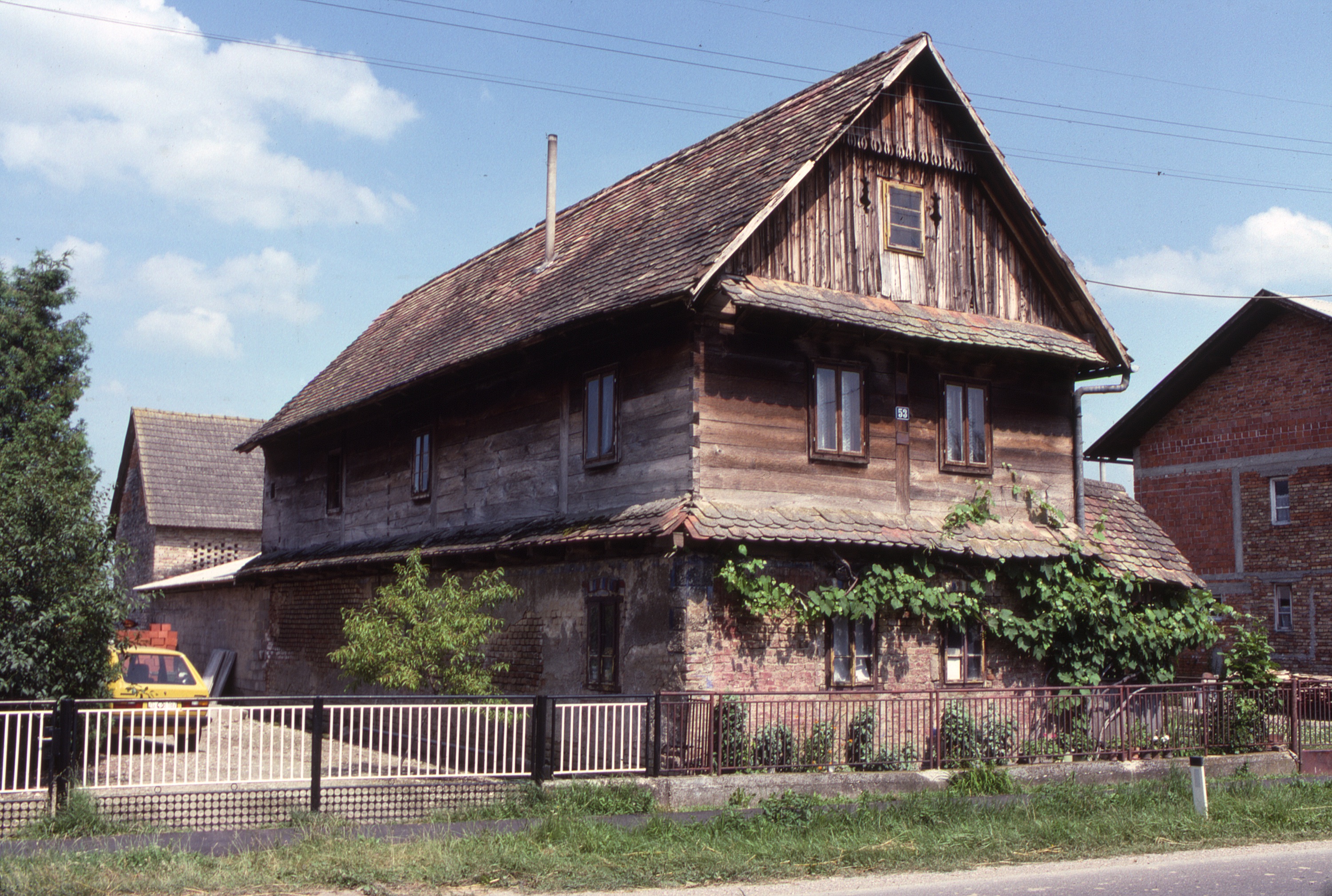 Judith Bing - <p>This wooden dwelling is referred to as a katnica or čardak in the region. Typically, these buildings have a gabled end and principal living spaces facing the street, and are constructed using oak planks with interlocking joinery at the corners. This čardak has been renovated with brick-faced walls at the ground level, which were originally constructed of exposed wooden planks and the interior spaces used principally to store equipment, livestock and produce. The renovated ground level is used as additional dwelling space. (photo 1988)</p>