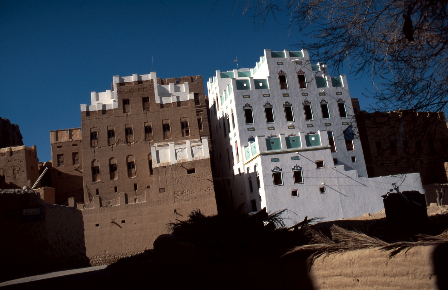 Peggy Crawford - Wadi Doan. A painted and a mostly unpainted multi-story mud brick building side by side