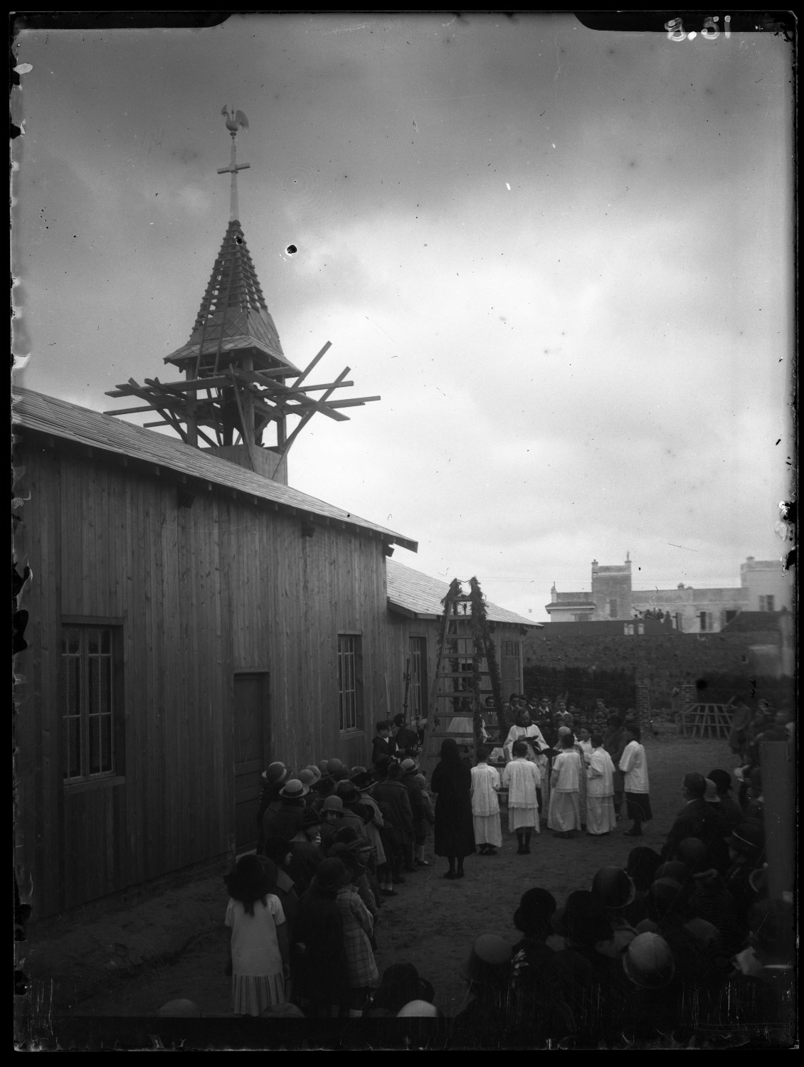 Crowd gathering around a building during construction of steeple on French religious school; steeple with cross and rooster