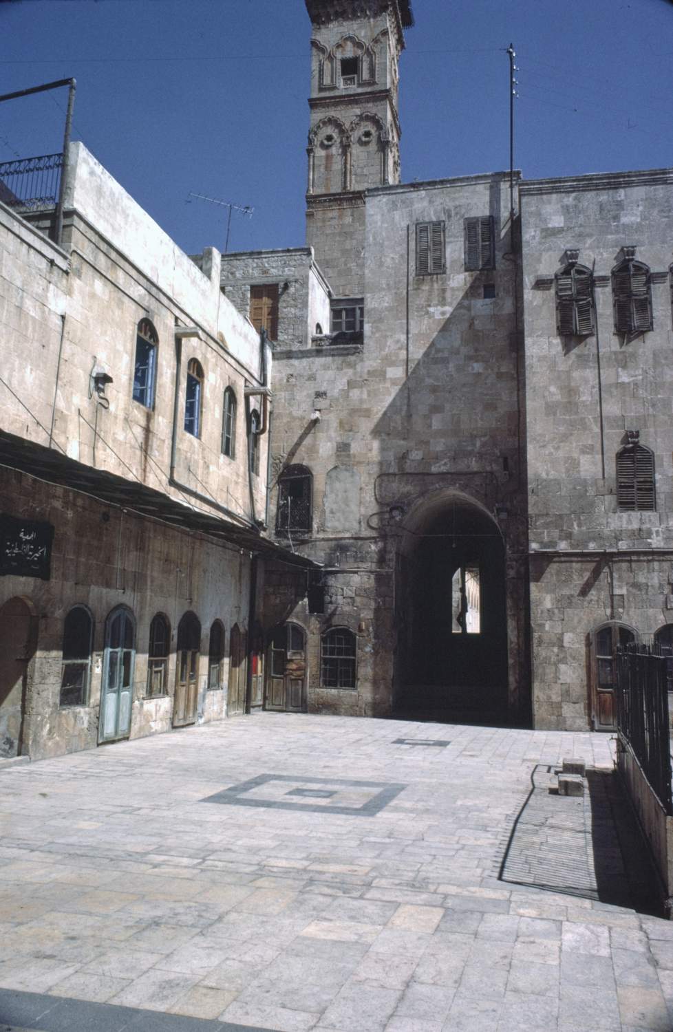 View of courtyard facing east showing archway of hall behind entrance portal in northeastern corner. Minaret of Umayyad Mosque visible in background.