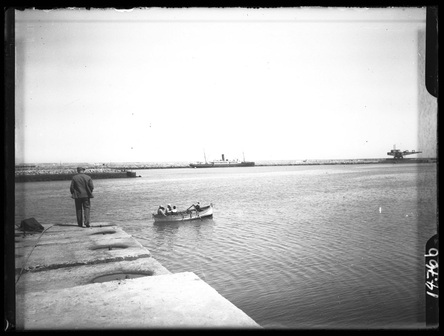 View of a man in European dress standing on the quay looking at the harbor as a rowboat passes