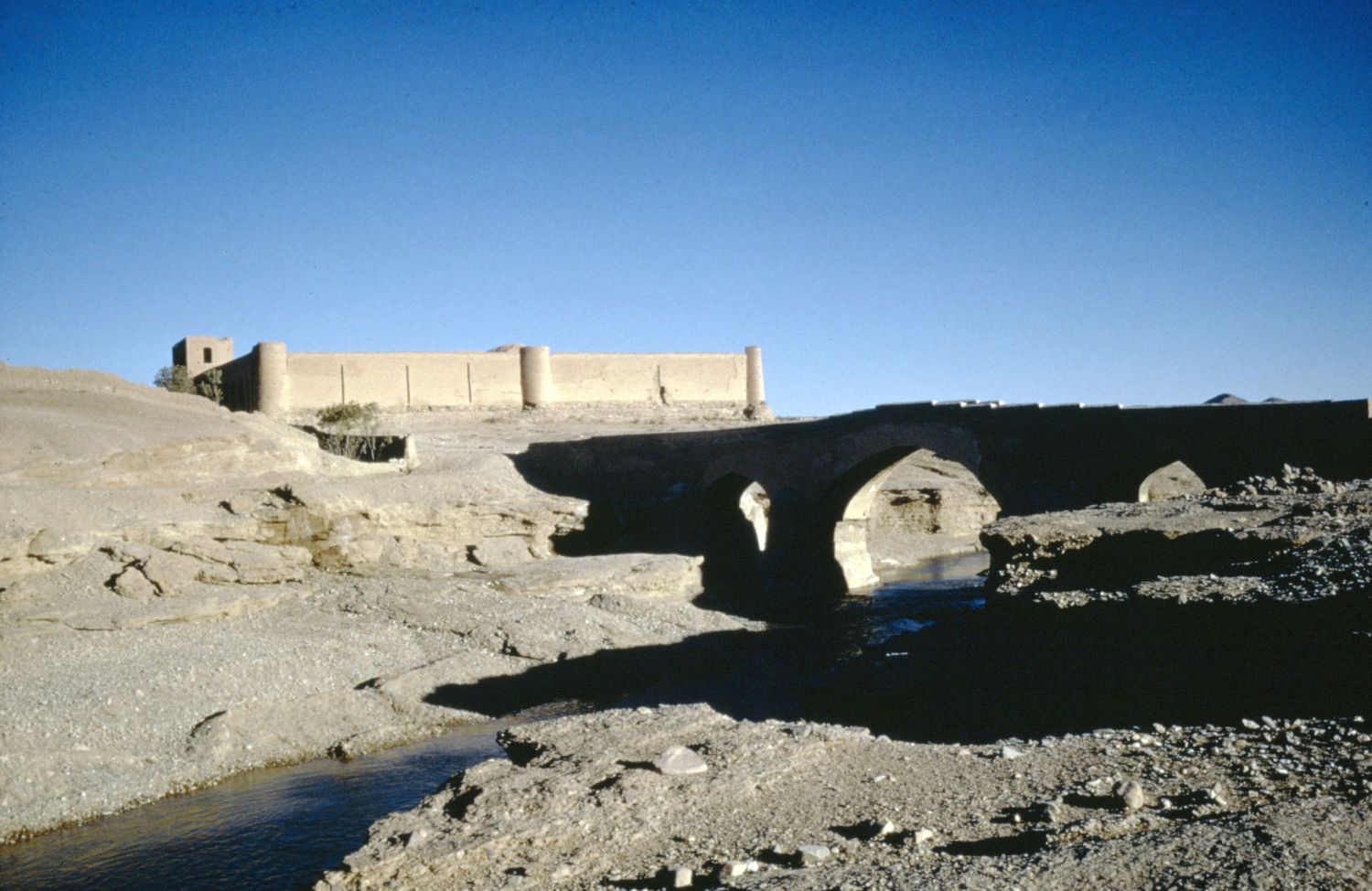 View of bridge over stream and walls of caravanserai in background.
