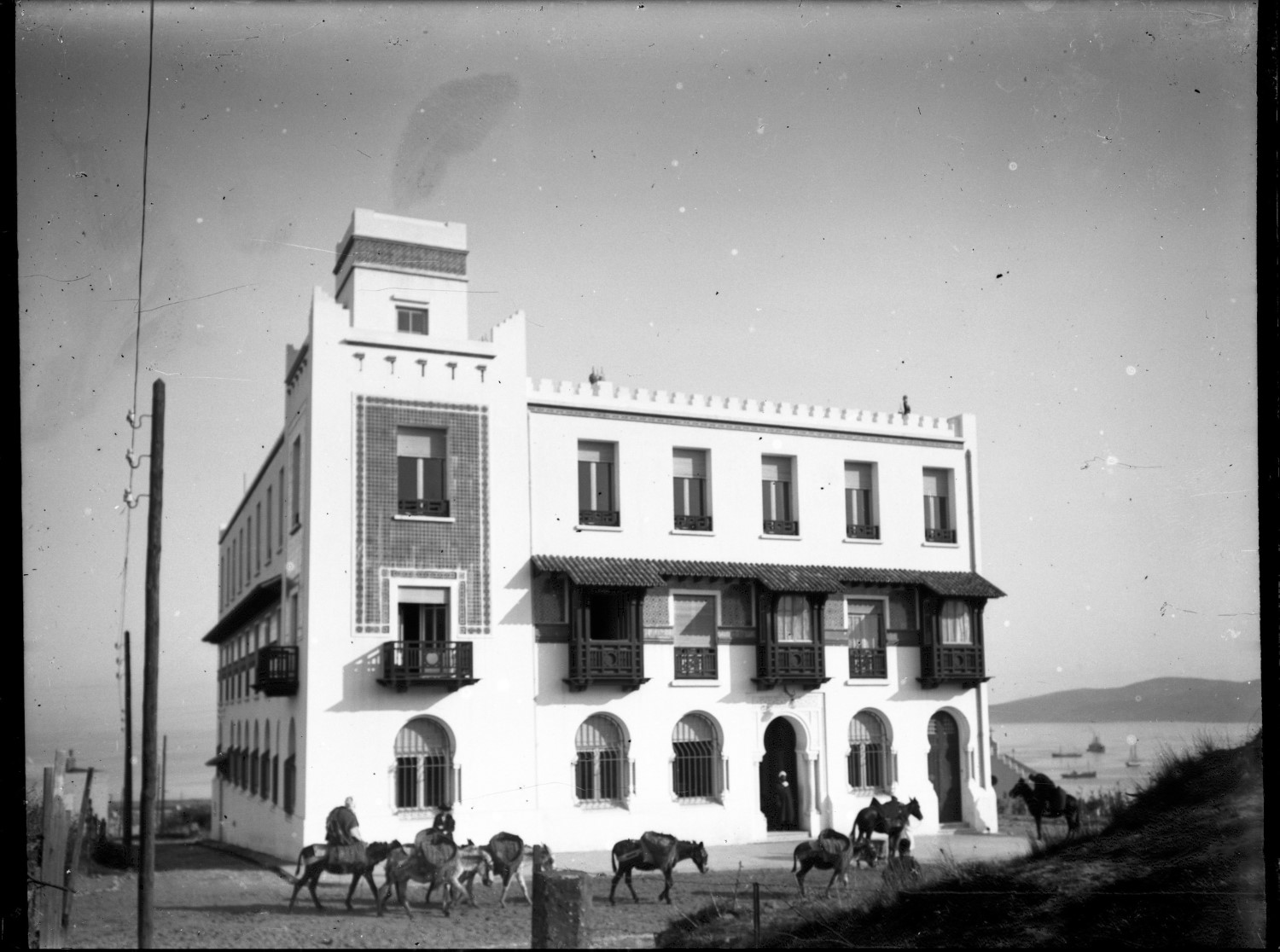 View of the facade with awnings, pack animals passing in front. A clear view toward the bay. 