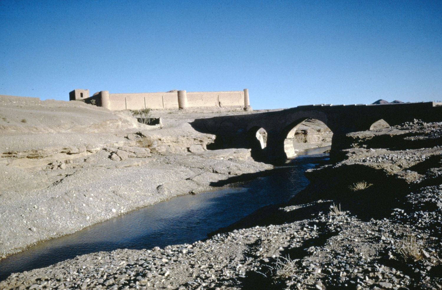 View of bridge over stream and walls of caravanserai in background.