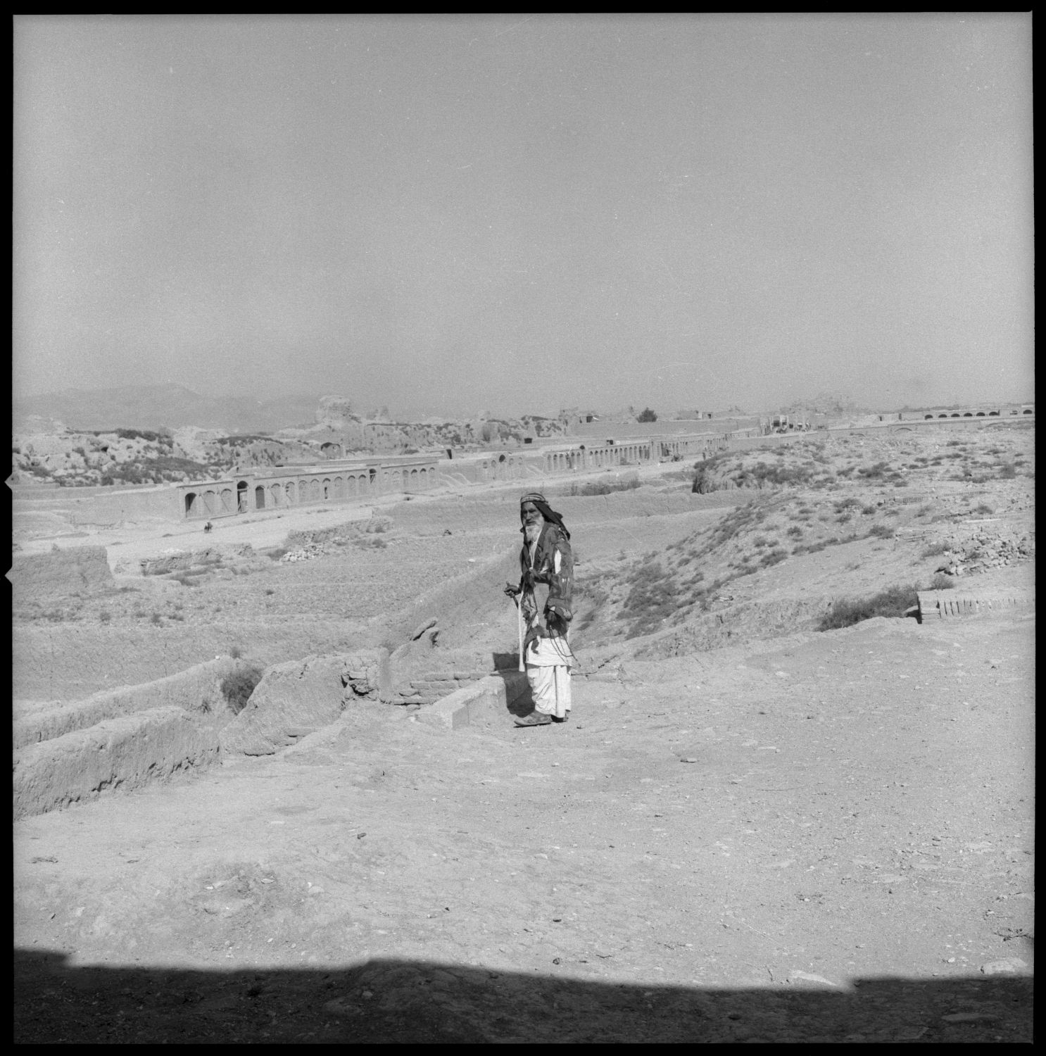 Man stands above dry riverbed with Pul-i Malan in distant background.