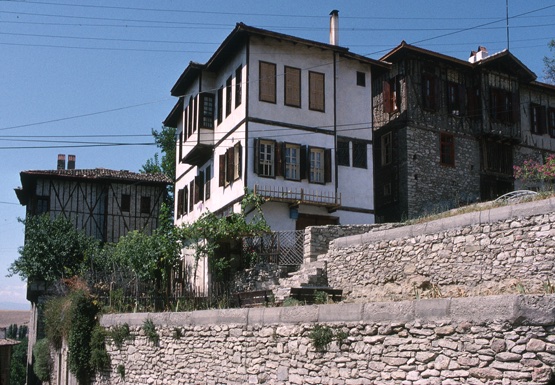 <p>These traditional houses in the Karaali Mahalla (neighborhood) date from the early 19th century. They were constructed of masonry, timber framing, soft lightweight infill brick, and clay tile roofs. The Direndefiller evi (house), in the center, is faced with a cementitious plaster coating. This view is looking southwest along Yokuşbaşi sokak. The house to the left is the Semerci Muharrem evi and is entered from Yokuşbaşi sokak (alley). The house on the right is a portion if the Tekeler evi that projects into the eski cami sokak, an upper alley to the northwest.</p>