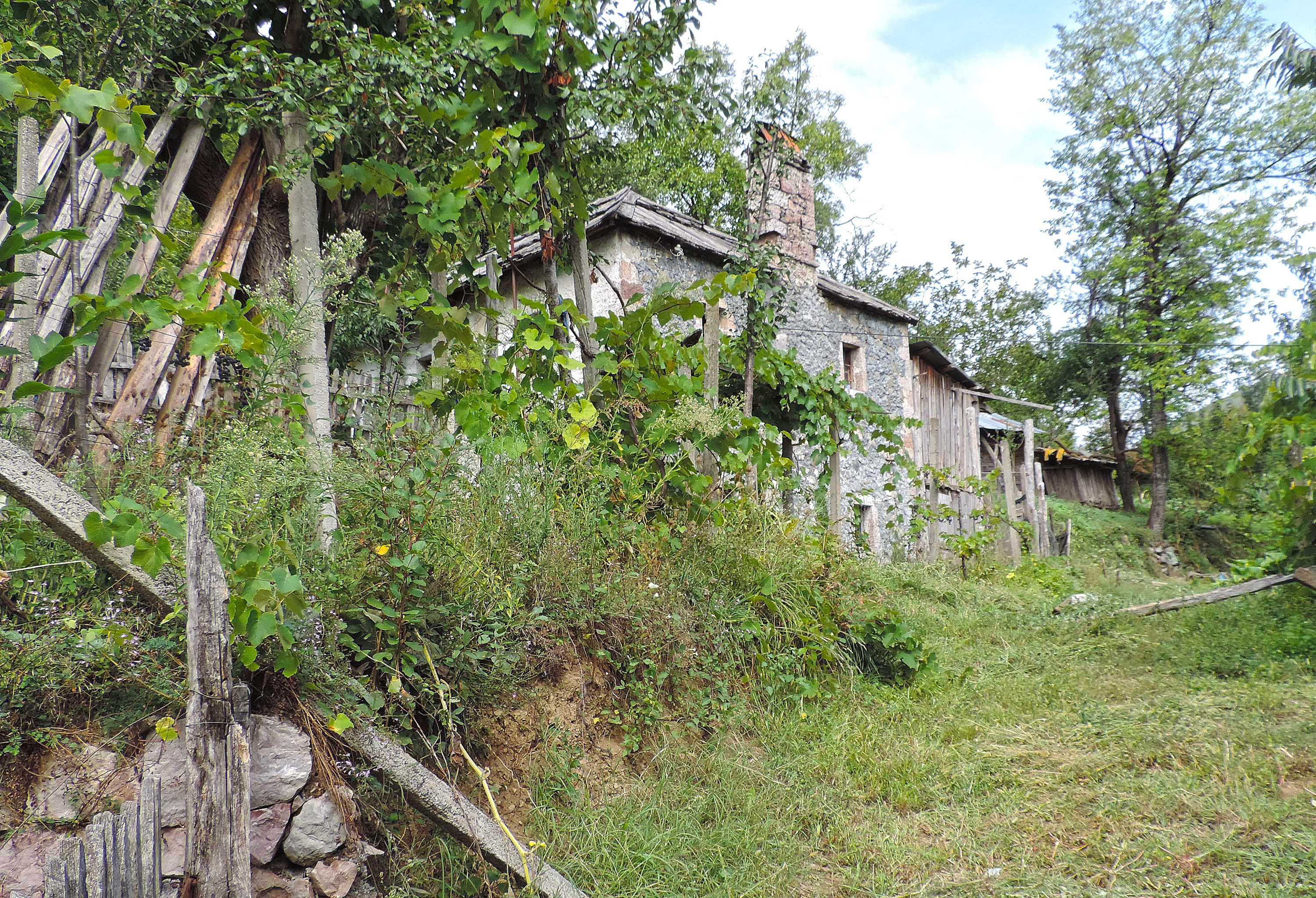 <p>This abandoned 2-story stone house is like a small kulla, square with peaked roof. A large chimney is flanked by window openings, and larger stones reinforce the openings and building corners. A wooden barn sits beyond the house.</p>