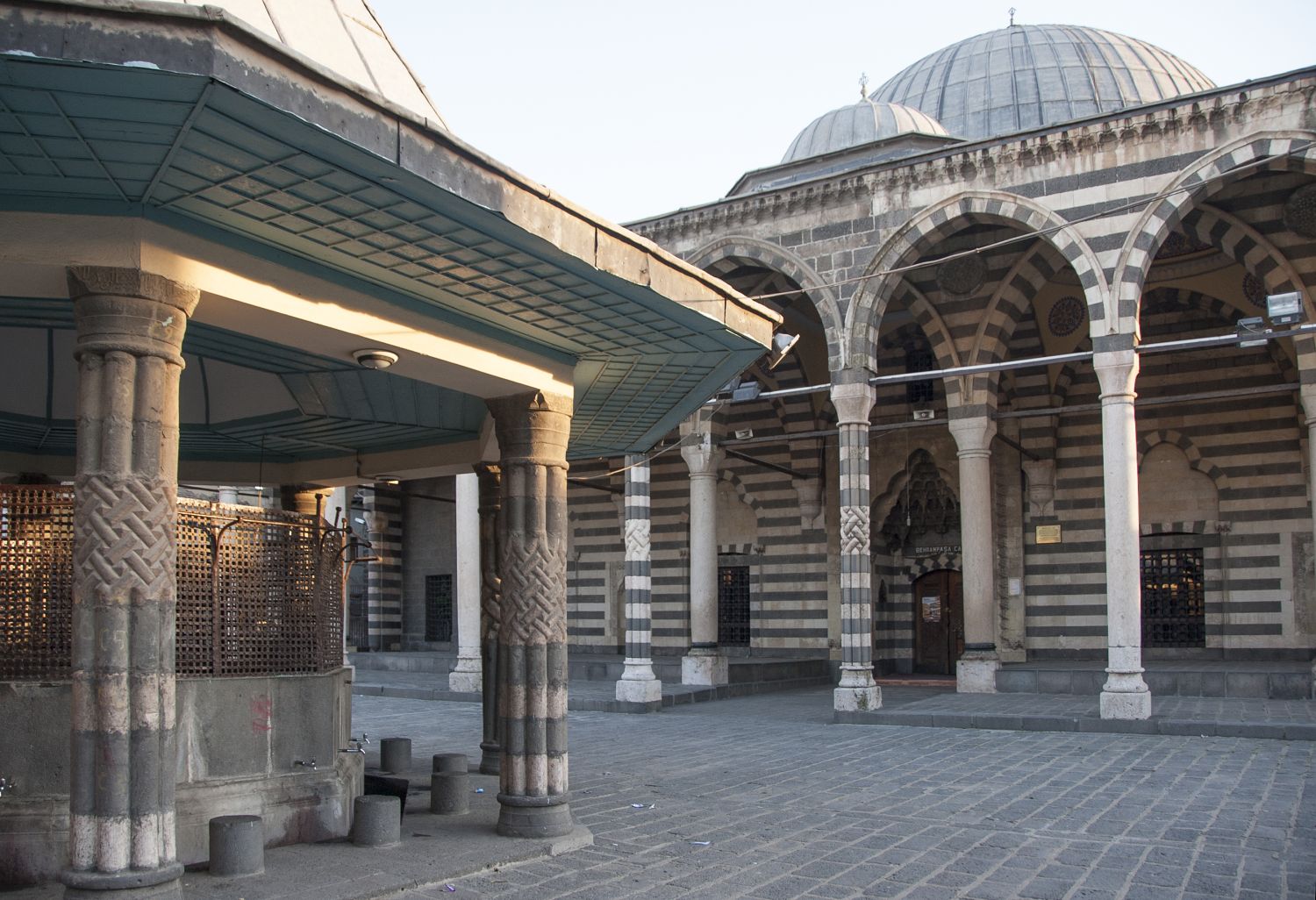 View of the paved square in front of the mosque. The ablutions fountain is visible in the foreground and the portico and north facade of mosque are visible behind.