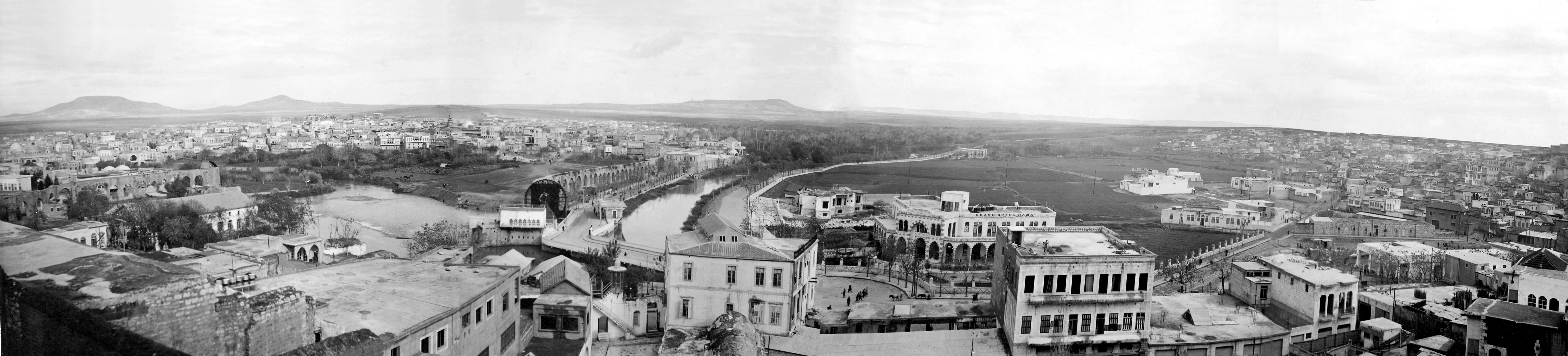  Hama - <p><strong>Hama, view from the Western Bank of Orontes River towards Al-Hadir Area</strong></p><p>Taken from the Al-Bashura quarter, this panorama captures the Al-Hadir area from the western bank of the Orontes River, where it curves northward. Notable features include the Noria al-Jisriyya and the water canal leading to the al-Arnaʾut palace. The scene also includes the new-looking hotel of Abi al-Fida and the al-Farraiya quarter on the right.</p>