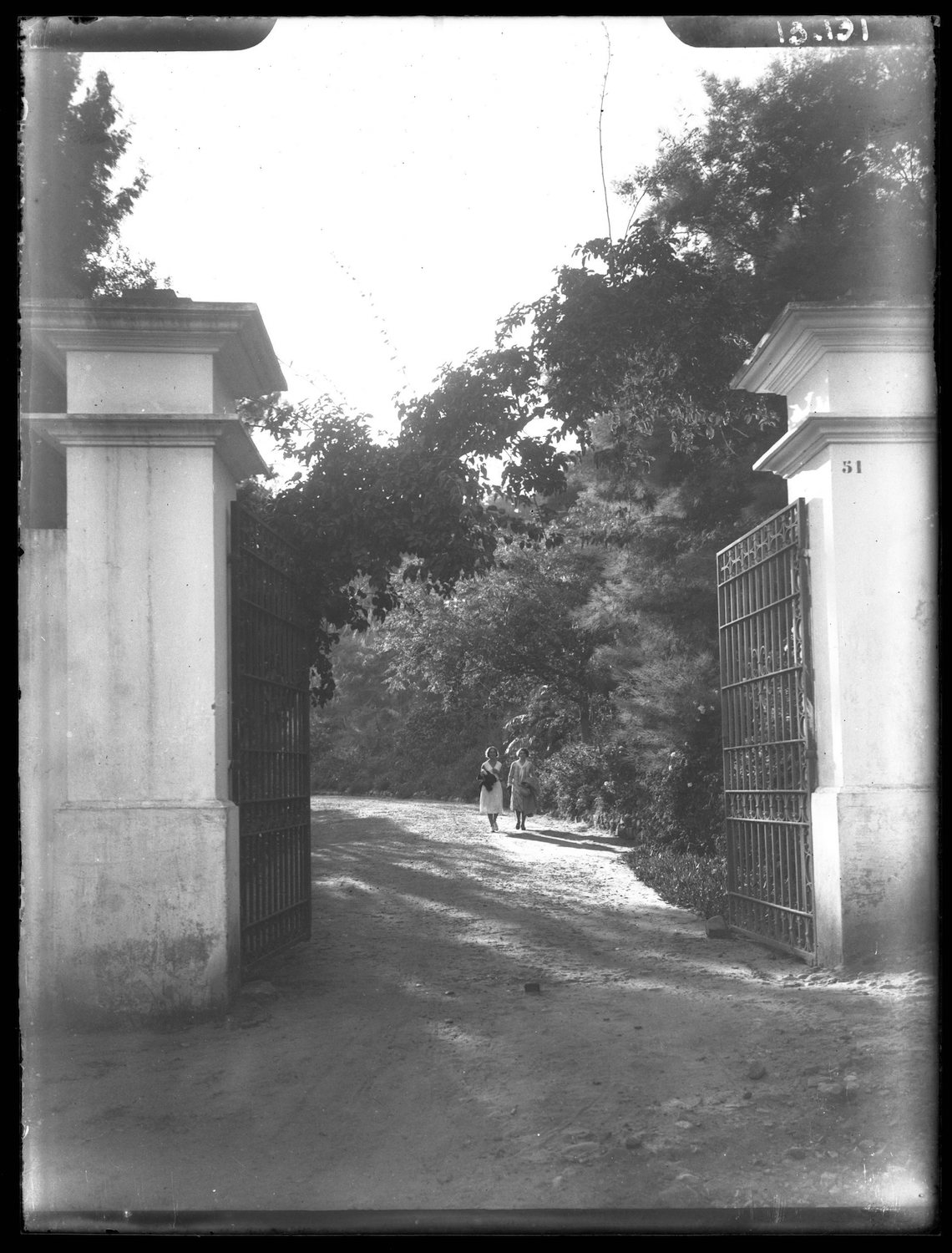 Two women in European dress walk towards the entrance gate of house number 51.