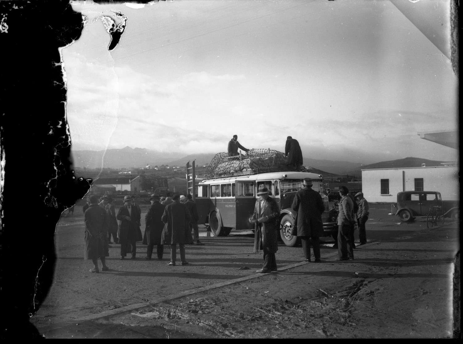 Gare de Oujda - Figures in European dress surround automobile being loaded by figures in Moroccan dress at the Gare de Oujda, with mountains visible in the background.