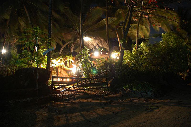 Exterior night view of the Madh Island house, showing 2 of the 3 lit barrel vaults and terrace canopy against the silhouette of a triangular gate