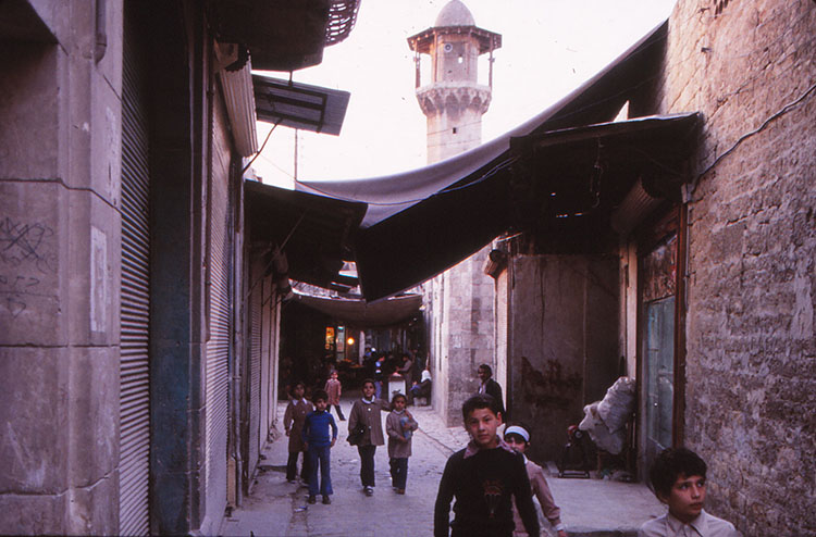 <p>View along a street in Bab Qinnasrin Quarter, Aleppo, showing awnings and minaret in background</p>