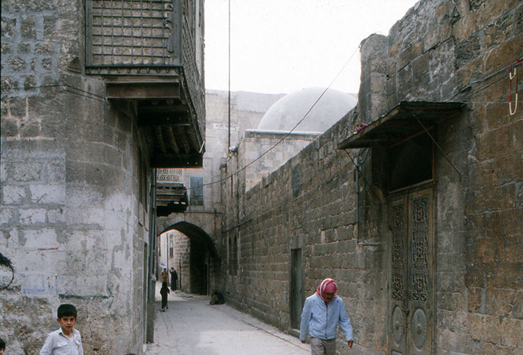 <p>View along street in Bab Qinnasrin Quarter, Aleppo, showing houses with projecting screened windows and arched bridge between buildings over street</p>