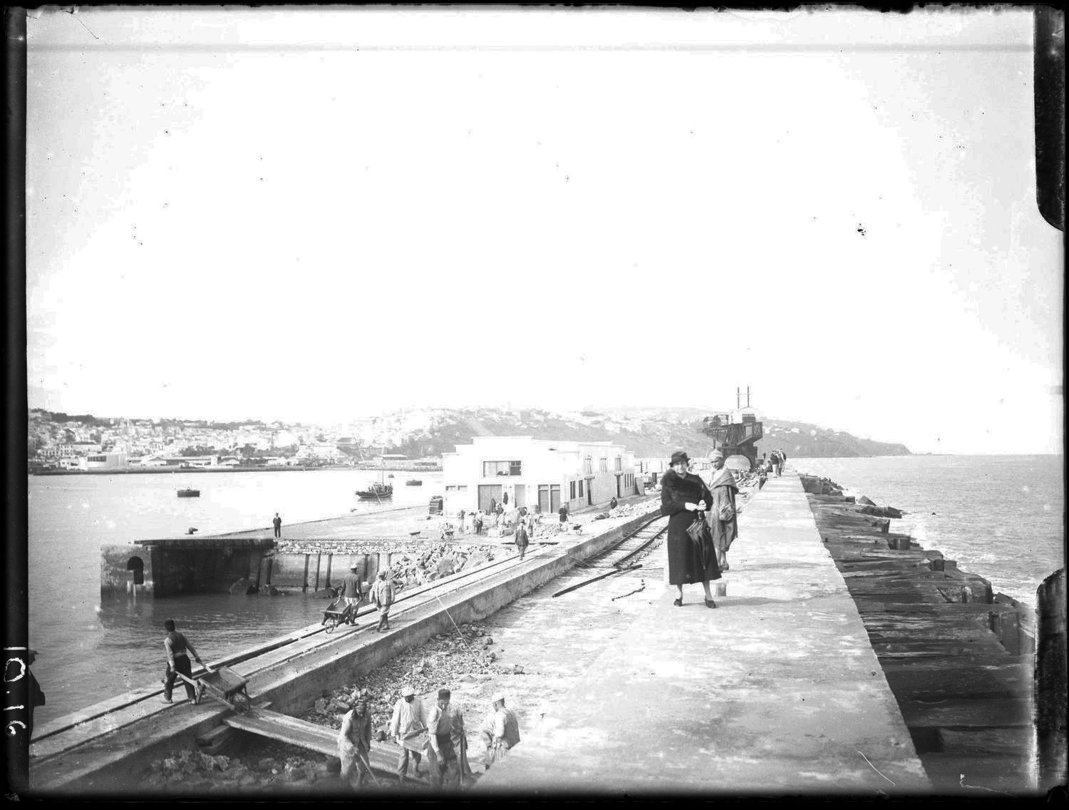 <p>View women in European dress on quai, construction men on left, city in background</p>