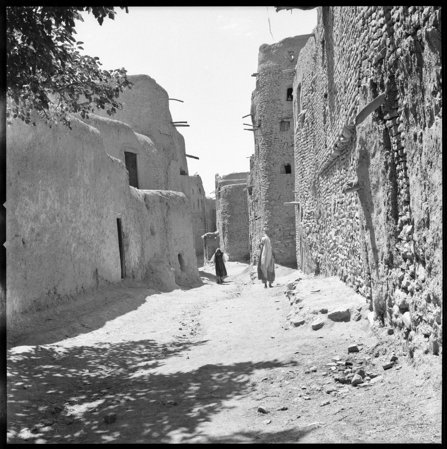 Pedestrians walk along narrow street between multi-story buildings with exposed roof poles in Ghazni, Afghanistan.