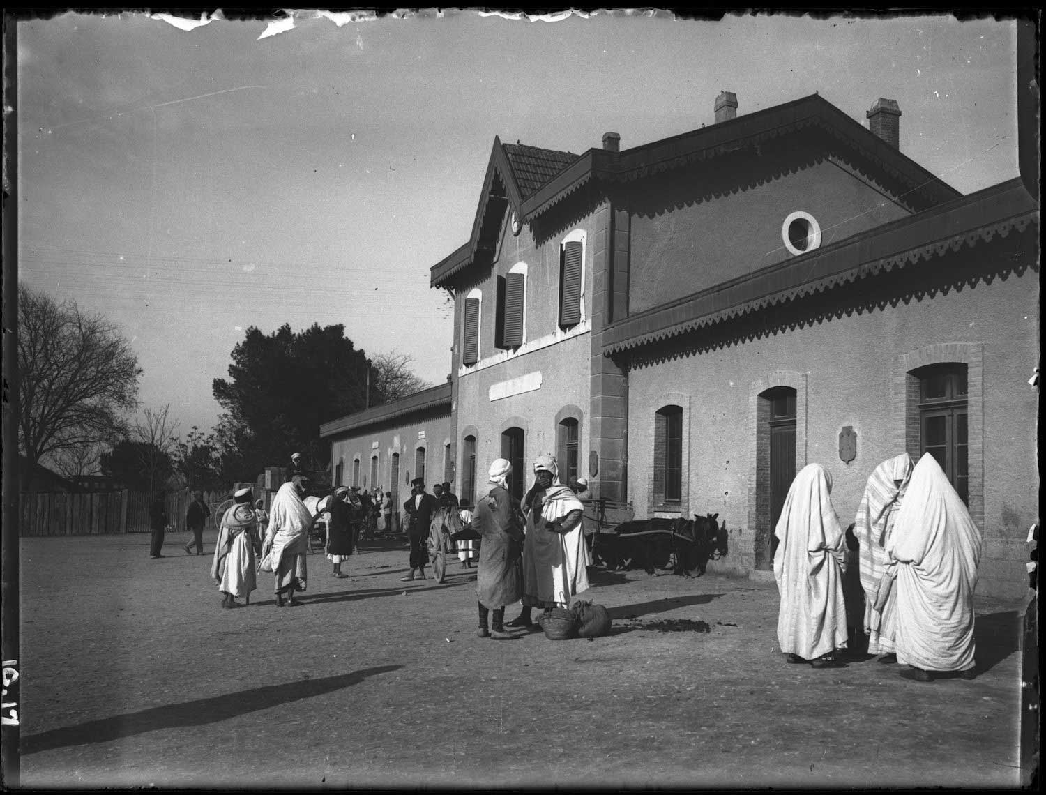 People, mostly in local clothing in front of a building, possibly a station