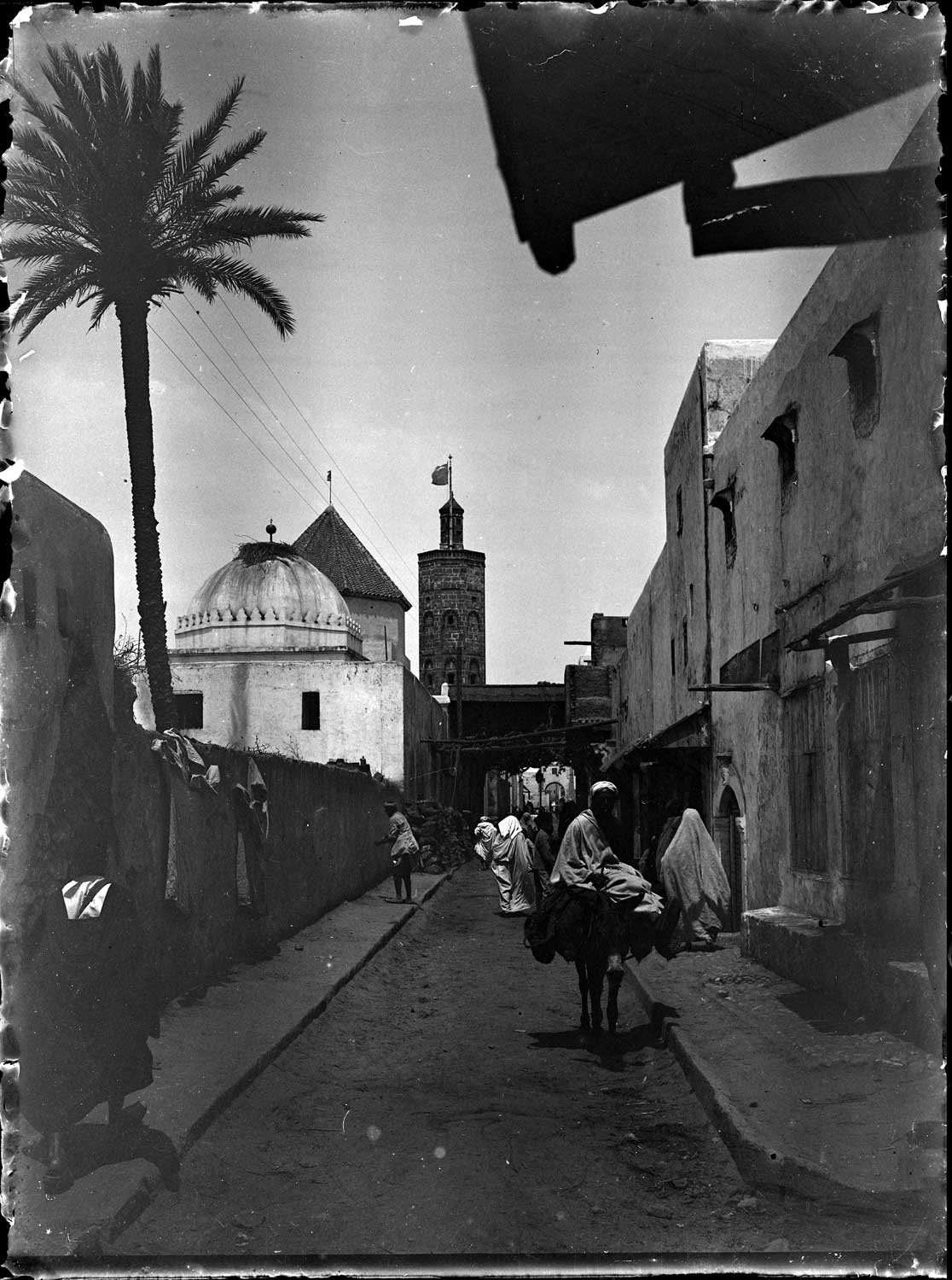 View down a street, a domed marabout, green tiled roof of a zawiyya, and octagonal minaret on the left