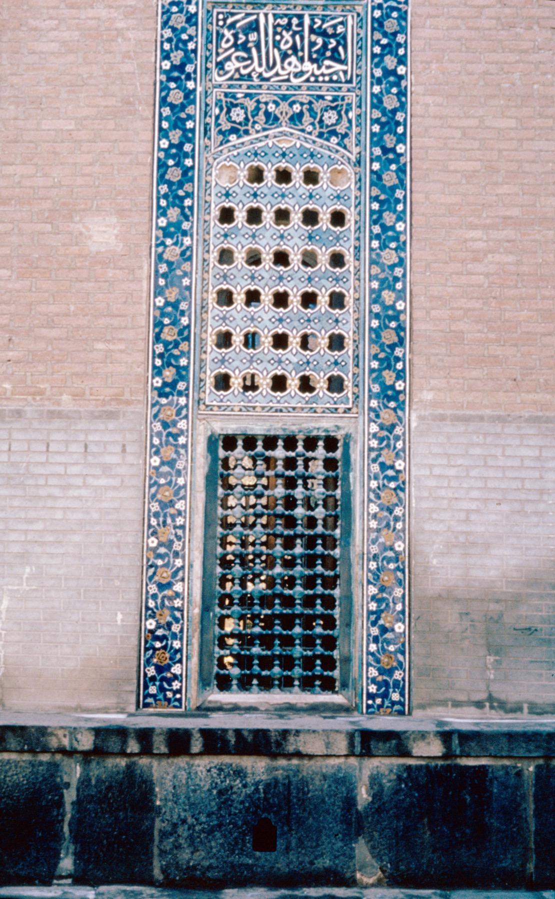 Exterior detail view of the courtyard wall of Dar al-Huffaz, showing screened window framed by tile mosaic band with floral motifs on a dark blue ground