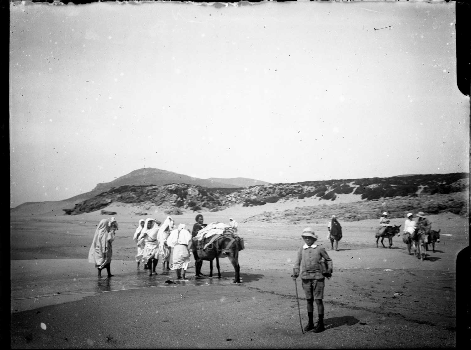 Moroccans with pack animals and a boy in European clothing on the beach of Achakkar (Cap Spartel)