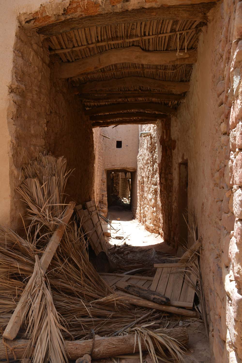 al-Ula - View down a covered passageway in old town with wood beam ceiling.  Palm fronds and wood on the ground.