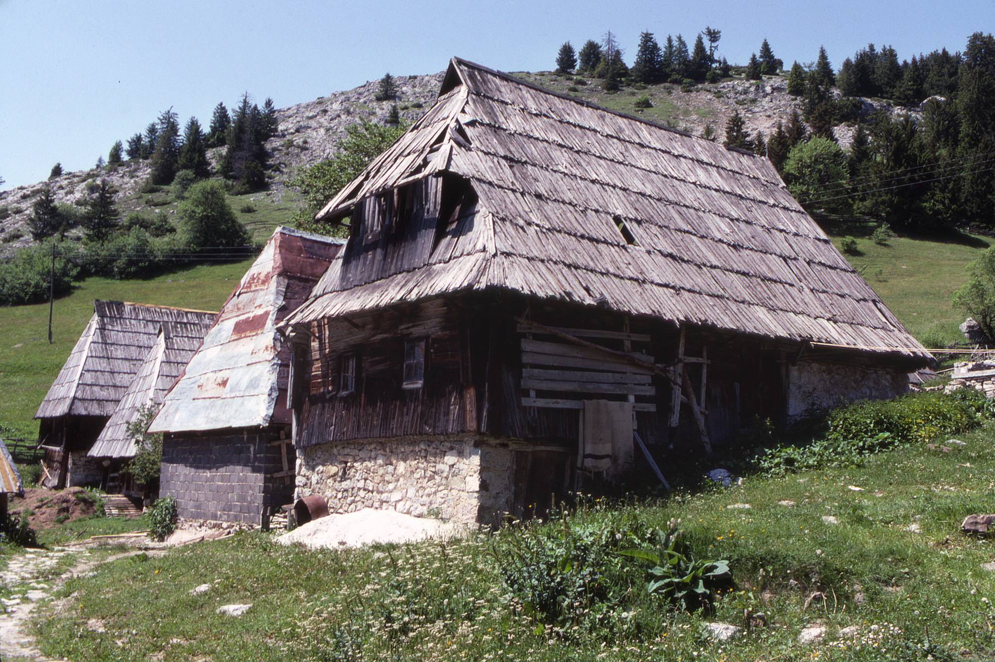 <p>An abandoned house with a roof-bound čardak space, with other structures, in this former sheep-herding village. The house upper level is primarily of wooden construction, including vertical plank wall siding, while the buildings beyond are largely of stone.</p>