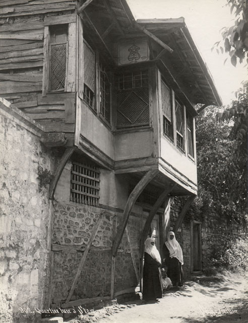 Wooden House on earthen street with rooms cantilevered over street wall and two women in street outfit standing before the entrance.  The garden entrance is seen further down the wall