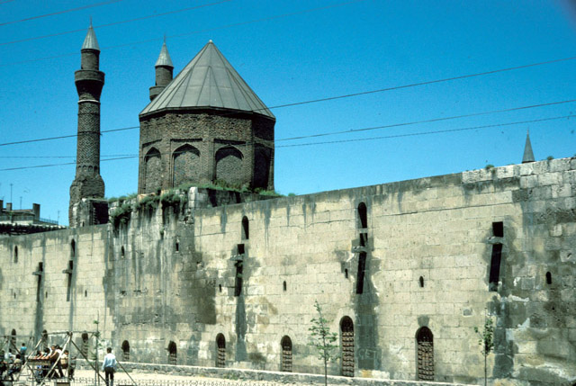 Exterior view from east-southeast showing southern wall and tomb tower, with the double minarets of the nearby Cifte Minaret Madrasa seen in the background