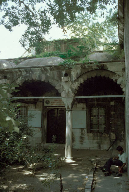 Exterior view of madrasa classroom, looking north, with madrasa wall seen on the right.  The three-bay portico preceding the classroom has white and red stone arches with rounded voussoirs and diamond-cut capitals; the wider central bay is crowned by a barrel vault
