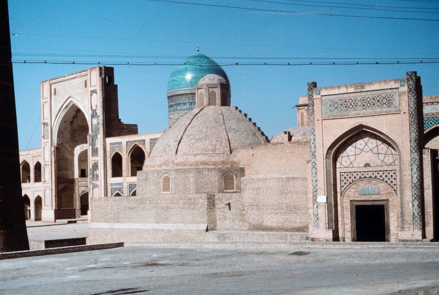 Amir Olimxon Madrasasi - Street view toward the plaza known as Pa-i Kalan. The entrance to the Madrasah-i Alim Khan is visible at right. The high pishtaq of the Madrasah-i Mir-i Arab and one of its turquoise domes is visible at left.&nbsp;