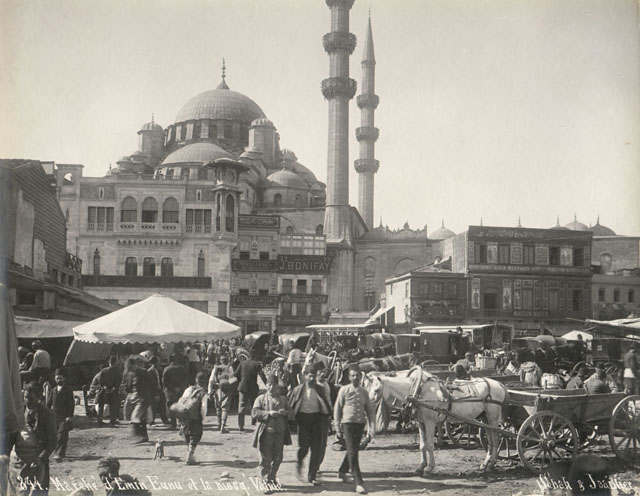 Yeni Camii Külliyesi - View of public square at the water's edge known as Eminönü Square, looking southwest. Framed with commercial structures adjoining the mosque, the square is seen crowded with pedestrians, horsecarts, horse-drawn streetcars and carriages
