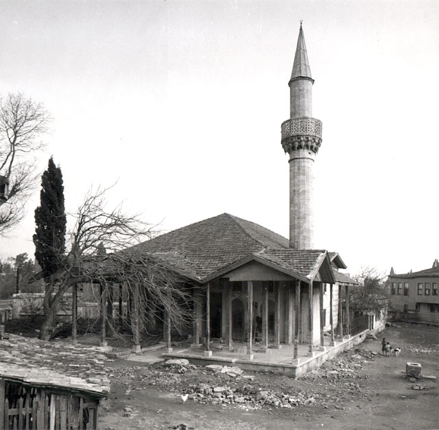 Exterior view from west-northwest, showing wooden columns and zigzag portico roof.  Seen immediately behind the mosque is the small mekteb and two-story wooden house
