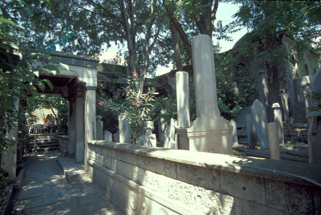 View looking north through gate on Yeniçeriler Street, showing pathway passing through tomb portico with eighteenth century cemetery to its right; the classroom and madrasa appear behind tombstones in the background