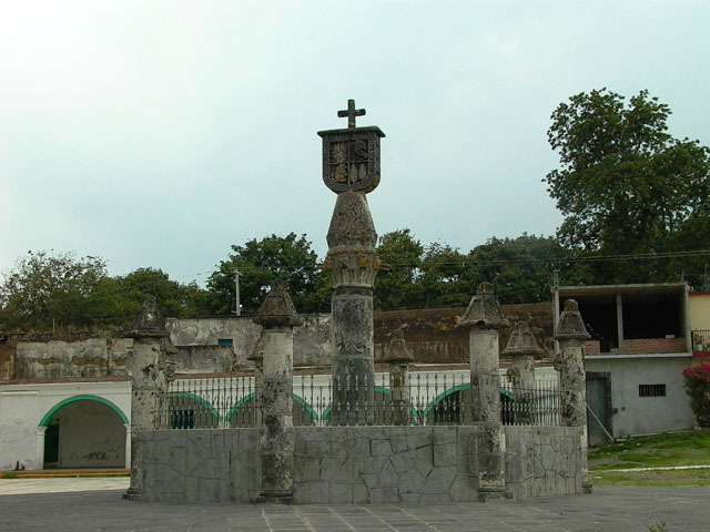General exterior view of stone fountain showing coat of arms atop a column, encircled by shorter columns and metal fence