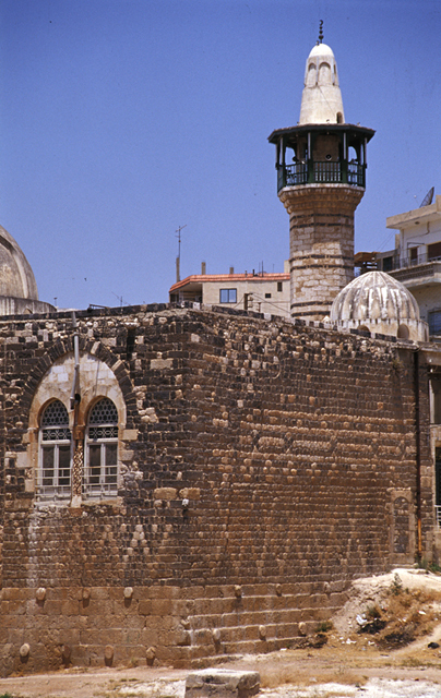 Partial exterior view from southeast, showing masonry construction -- calcarous ashlar foundation with antique spolia and basaltic stone walls, a prayer hall window, minaret, and tomb dome (left)