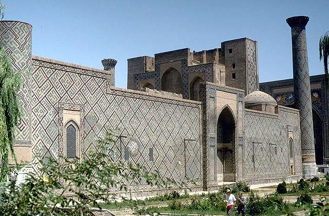 Exterior view showing the south elevation with two minarets at the corners and an arched portal opening to the courtyard. The Shir Dar madrasa is visible in the background