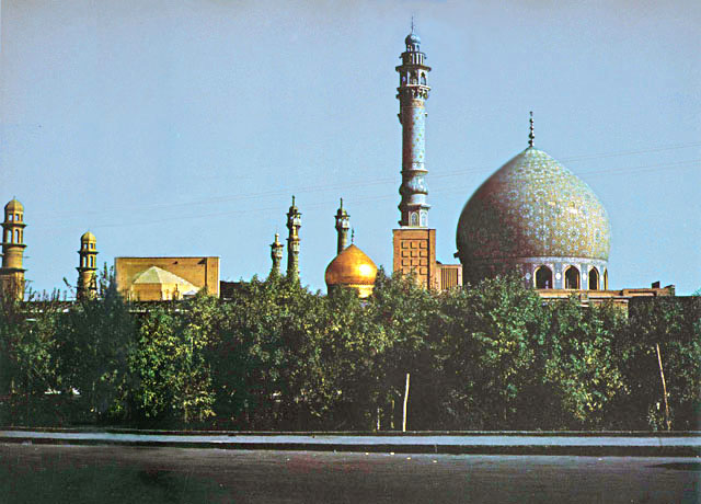 Exterior view from the Qum river, looking east at mosque dome and portal. The golden dome of the Hazrat-i Ma'suma Tomb and twin minarets of the Mirror Iwan appear in the middleground