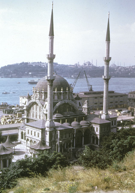 Elevated view from north-northwest, showing mosque with tent-like roofs of the sabil, the timekeeeper's room and the ablution fountain seen in the courtyard to the left and trade entrepots behind the mosque. The silhouette of the historic peninsula appears beyond the Golden Horn in the background