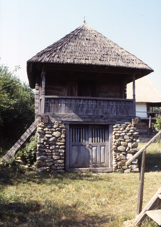 <p>This service structure from the village of Cârligei has a stone base and interlocking timber plank upper level. The building sits next to the 19th century dwelling within the same gospodărie, or homestead. The ground level is a storage cellar, and the upper serves for clothing storage, and has a porch (privdor) for outdoor activities. The wedge shaped wooden shingles appear to have been formed to take advantage of the natural splitting planes of the wood.</p>