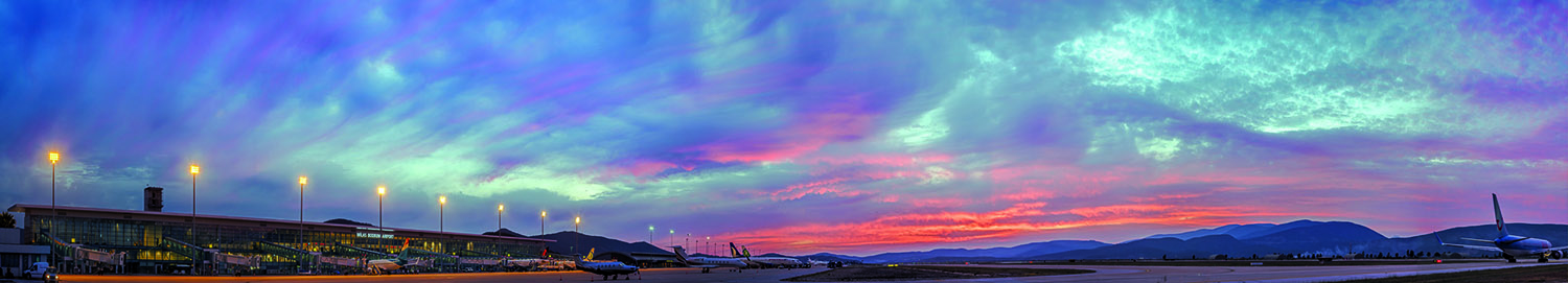 <p>Panoramic view at dusk of the airport terminals facade and bridges, along with the runways and the scenery of the meadows up to mountains</p>