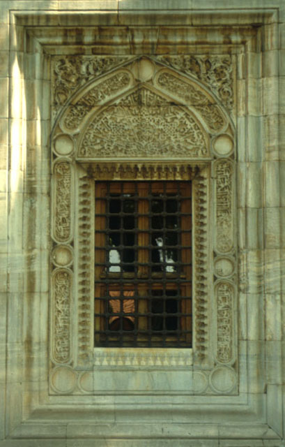 Exterior detail of a window protected with iron screen and framed in marble saw-toothing, scriptures, interlocking floral patterns and moulding creating a three-dimensional composition
