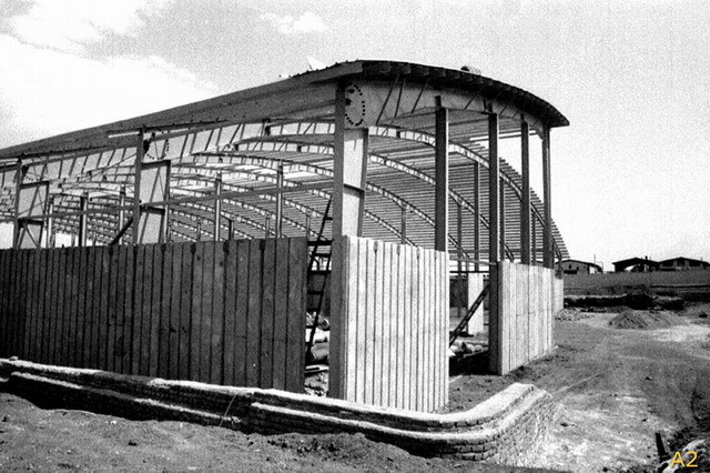 Exterior view, during construction, showing curved steel structure and concrete wall panels
