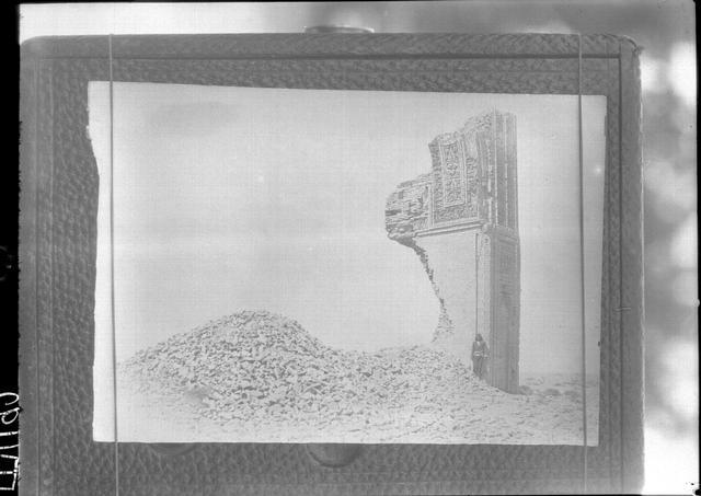 Photograph, pinned to a board, of one half of a standing ruined arch with a heap of collapsed bricks in front