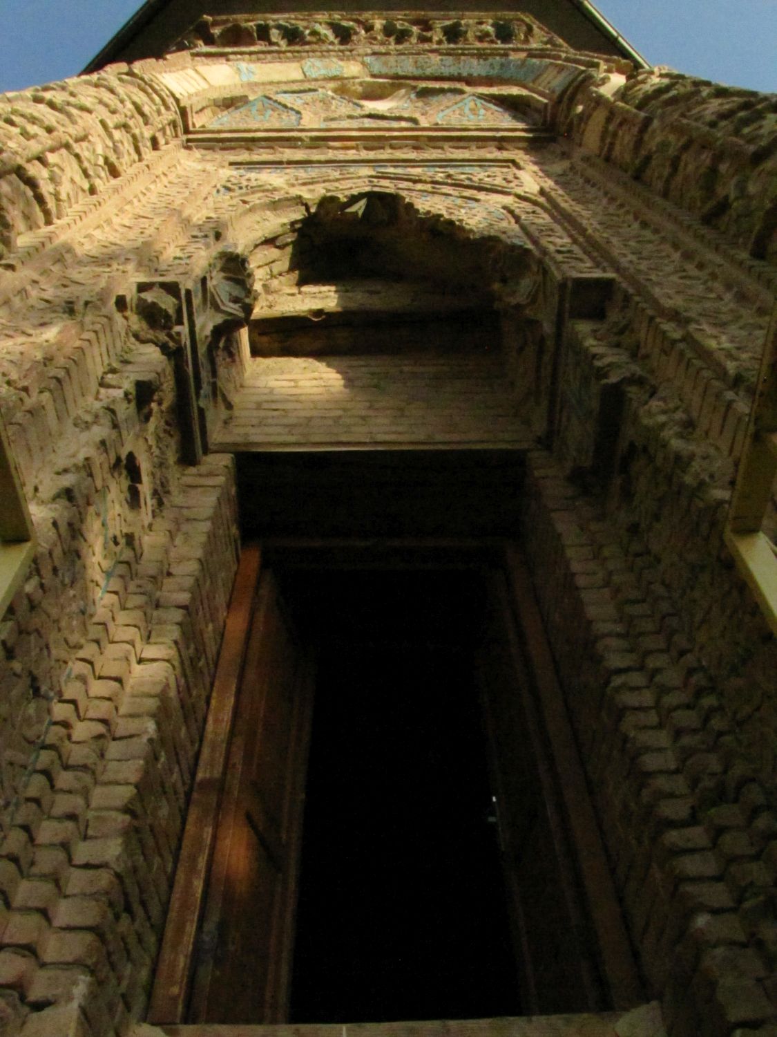 Exterior view up at doorway and top of portal, with damaged muqarnas, partially glazed tiles and brickwork