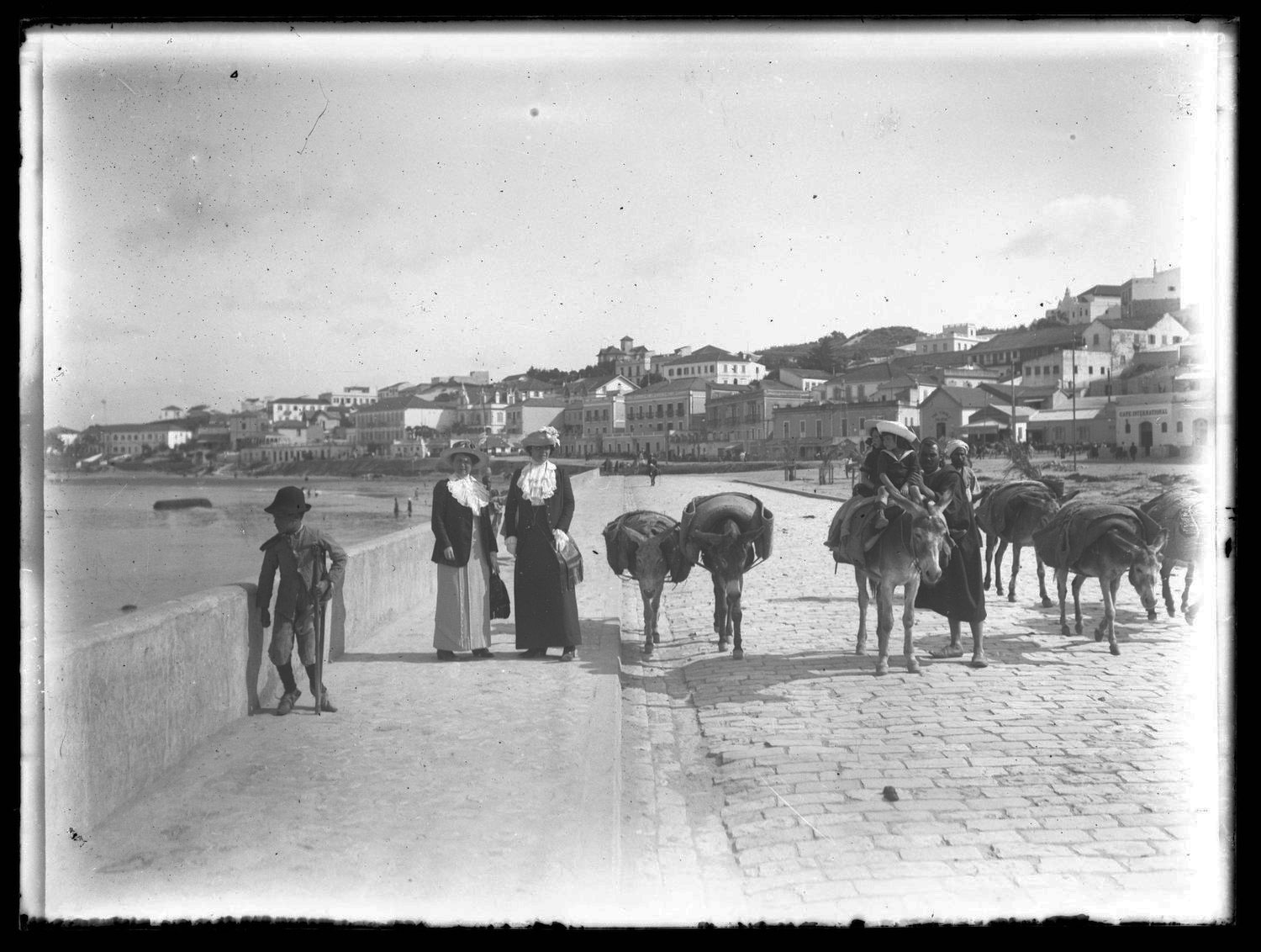 View of a boy walking with a crutch and two European women posing alongside the beachfront boulevard where several mules, one carrying two children, and their Moroccan attendant are approaching