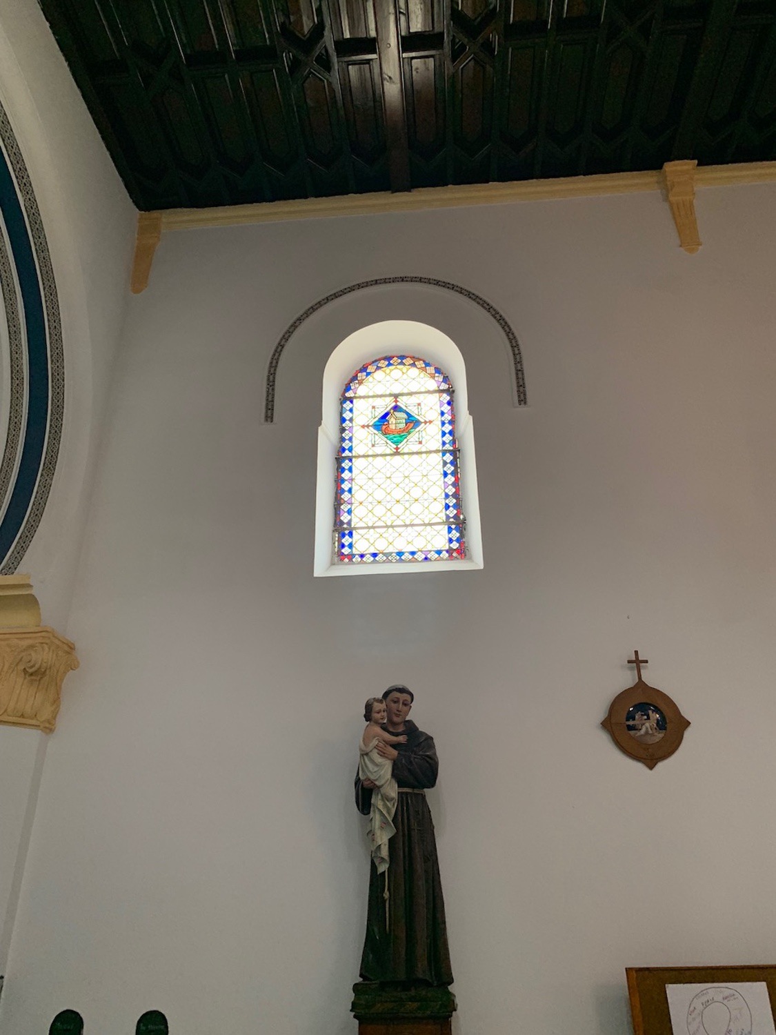 <p>Interior view a stained glass window, under which is a statue of Saint Anthony</p>