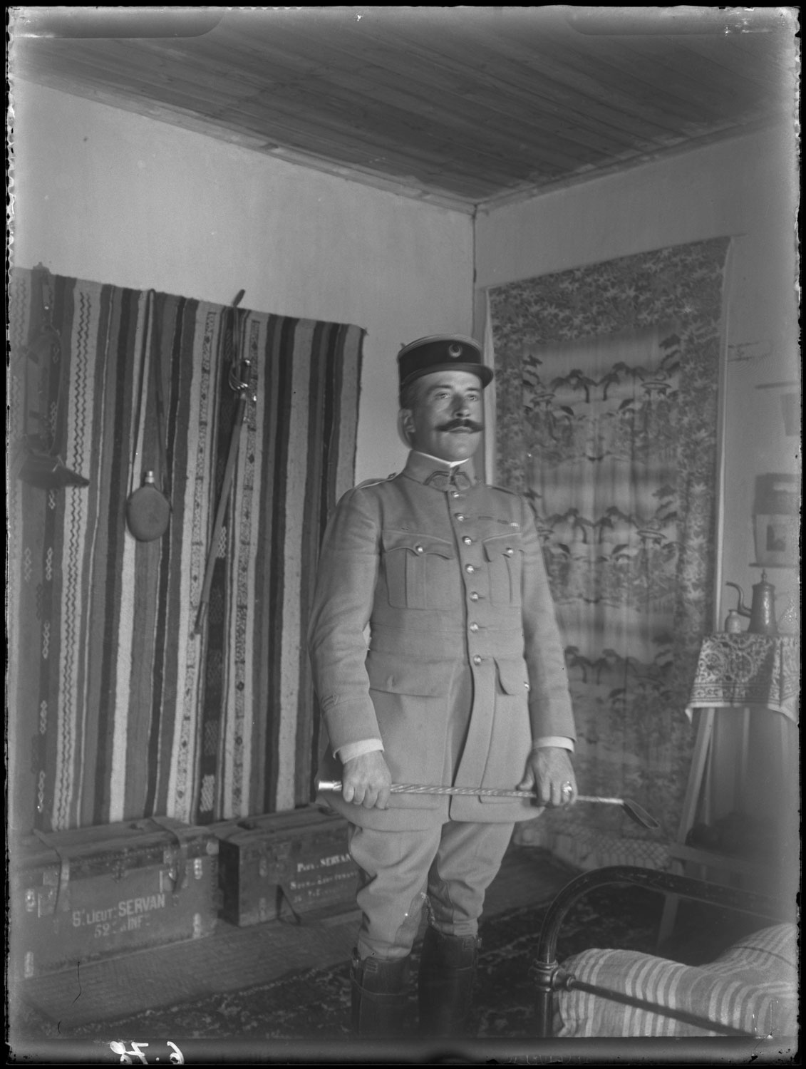 Interior portrait of a soldier at attention, textiles decorate the walls. Probably Paul Servant. 