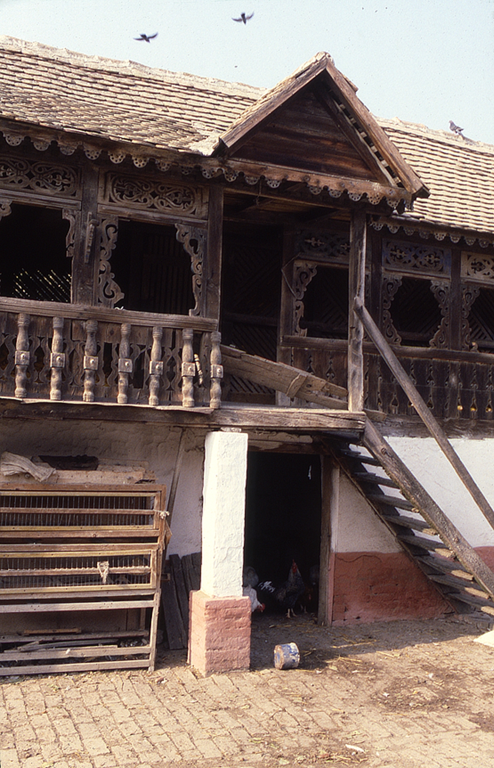 <p>The wooden rail at the stair landing is now missing but the notched corner post indicates where the railing cap once existed. On the right, more solid wooden railing panels occur and the openings above reveal the koš lattice work set back from the surface. The extended cross gable that covers the landing serves as a boundary for the gently sloping tile roof over the gallery.</p>