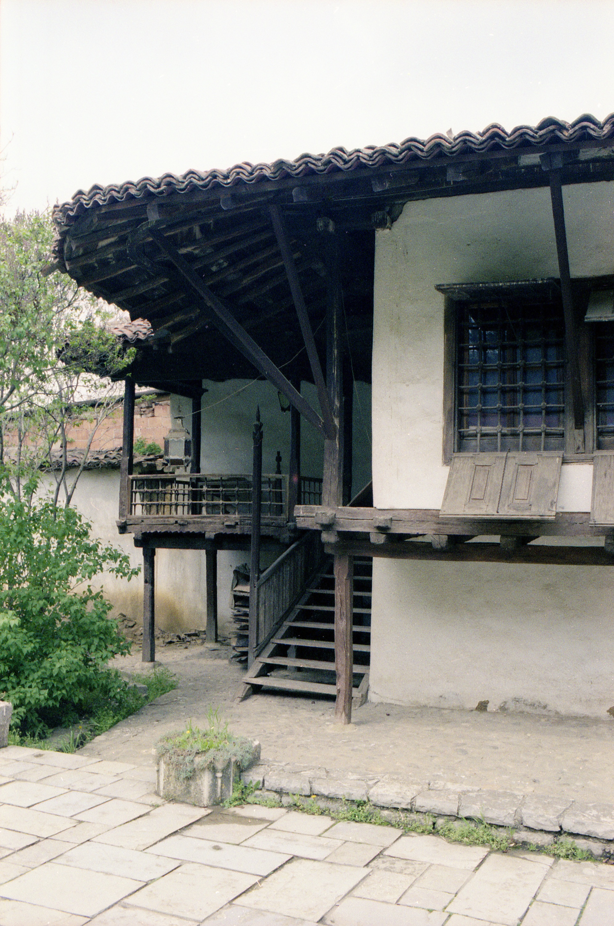 <p>This 18th century house, with its čardak (raised porch) sits within a walled garden to which it was relocated as part of the Ethnological Museum in Priština. In 1991 the interior cabinetry and some built-in furniture was in place along with artifacts of the period when it was built. One can see, under the stairway, timber for ongoing work. The broad roof projections are supported by pronounced brackets that connect to posts rather than to the wall. The interlocking beams, supporting the čardak deck, contain decorative notched patterns.</p>