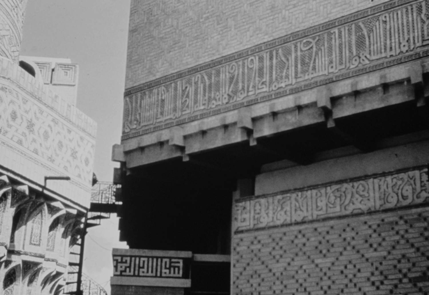 Detail view showing brickwork, geometric patterns, and Kufic inscriptions on the exterior of the prayer hall, and partial view of the base of the restored minaret.